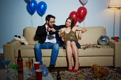 Middle-aged Asian man flirting with pretty young woman and pouring champagne into her flute, she enjoying night while sitting on couch in living room