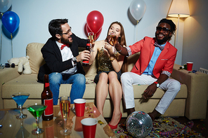 Pretty young lady in stylish clothes sitting with her male friends in living room and clinking glasses together