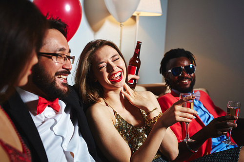 Portrait of cheerful young woman with red lipstick holding champagne flute in hand and laughing at funny story told by her Asian friend