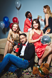 Well-dressed colleagues with champagne flutes and beer bottle posing for photography while Afro-American man and blond-haired woman standing behind them and clinking glasses together