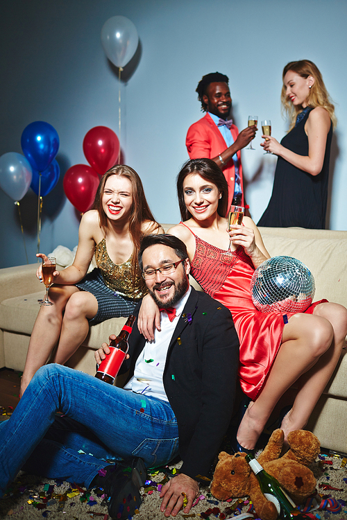 Attractive young women with champagne flutes and Asian man with beer bottle posing for photography on foreground while multiethnic couple toasting on background