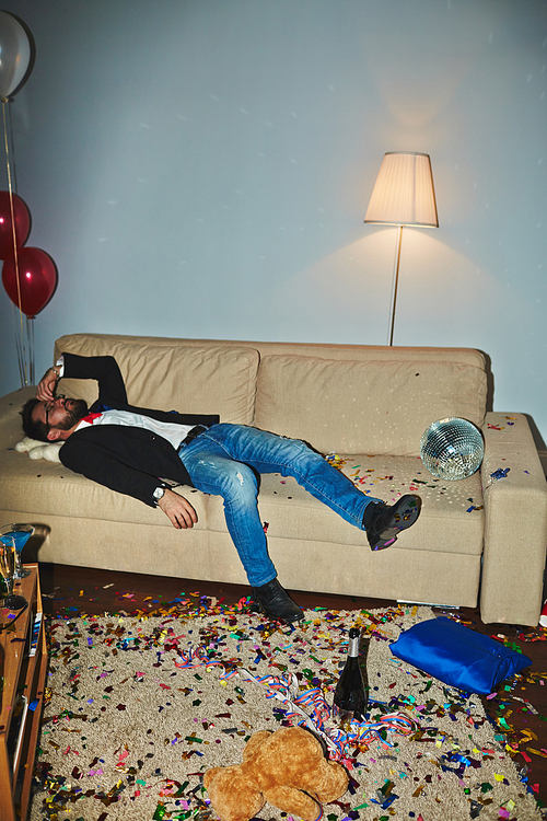 Messy room after wild party: colorful confetti thrown everywhere, empty alcohol bottles and champagne flutes standing on floor and coffee table, Asian man sleeping on couch