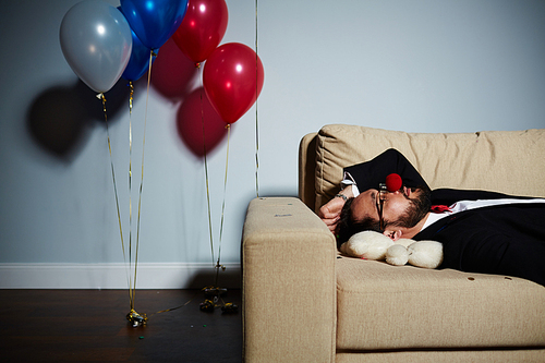 Waist-up portrait of bearded man with red clown nose sleeping on sofa after wild party, white bear lying under his head