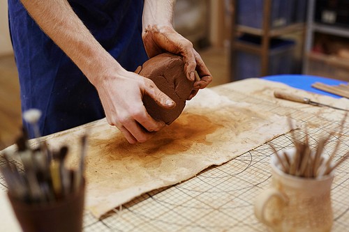 Hands of master kneading brown clay over workplace