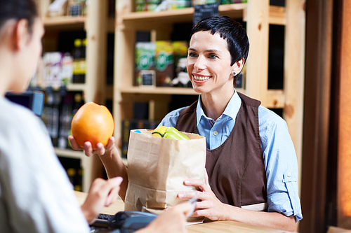 Waist-up portrait of friendly cashier putting fruits in paper bag while female customer paying for shopping