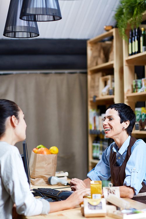 Profile view of middle-aged shop assistant and young customer having small talk while standing at cashdesk full of organic products