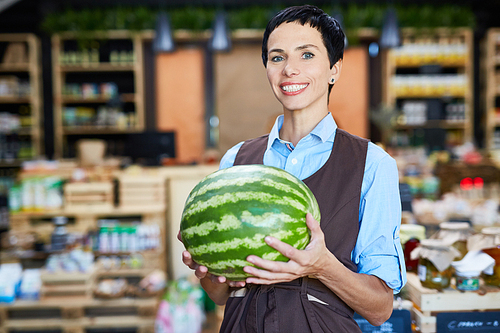 Portrait of cheerful shop owner in apron  while holding watermelon in hands, lovely store of organic products on background