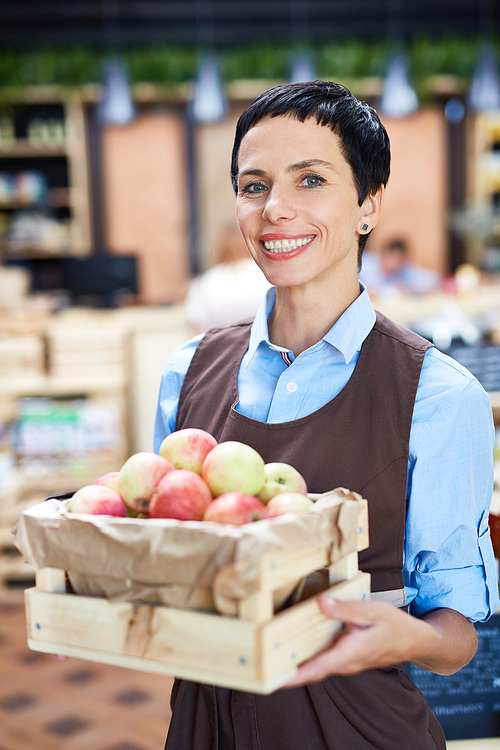 Black-haired store owner holding small wooden crate with ripe apples in hands while posing for photography, blurred background