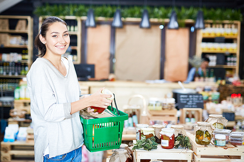 Portrait of attractive young customer with shopping basket  while holding jar with pickled tomatoes in hands