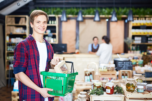 Doing shopping in supermarket: smiling male customer posing for photography while holding full shopping basket in hands