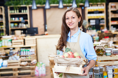 Waist-up portrait of pretty young shop assistant holding wooden crate with ripe apples in hands while  with charming smile, lovely store with organic product on background