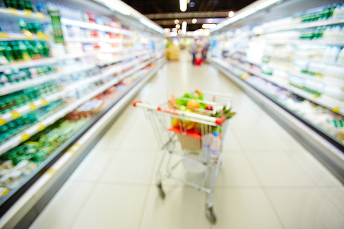 Blurred shopping trolley full of water bottles, fruits and vegetables standing between two refrigerators with dairy products