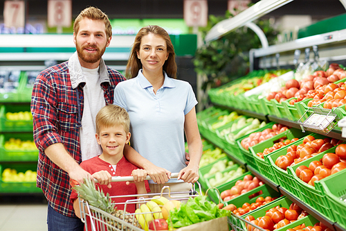 Cheerful family of three with full shopping trolley posing for camera in fruit and vegetable department of supermarket