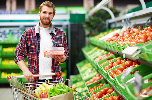 Young bearded bachelor holding packaging box with cherry tomatoes and  while shopping in fruit and vegetable department of hypermarket