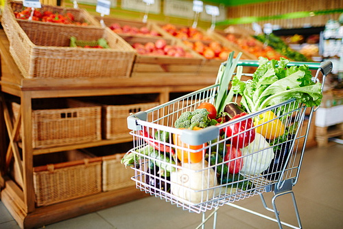 Shopping cart with variety of fresh ripe vegetables in modern hypermarket