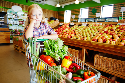 Adorable buyer pushing shopping cart in modern hypermarket in vegetable department