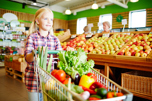 Cute little girl pushing shopping cart with fresh vegetables in supermarket