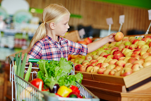 Adorable girl with shopping cart taking apple while choosing fruit