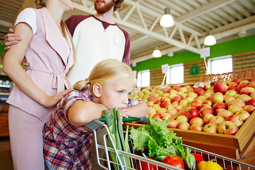 Tired girl leaning on shopping cart with her parents behind in supermarket