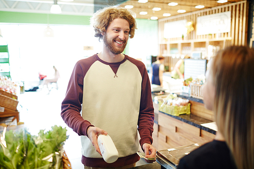 Happy young man showing bottle of milk to cashier and going to pay by cash