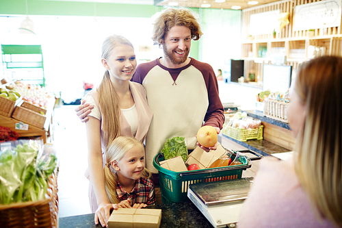 Young family of three standing by cash-desk with basket full of products