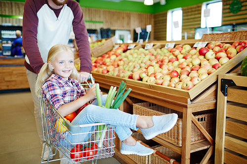 Happy girl sitting in shopping cart while her father pushing it while choosing food products in supermarket