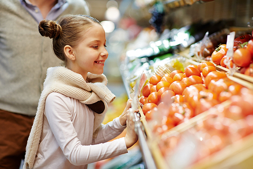 Side view portrait of cute little girl smiling leaning over vegetable counter with fresh ripe tomatoes and other vegetables