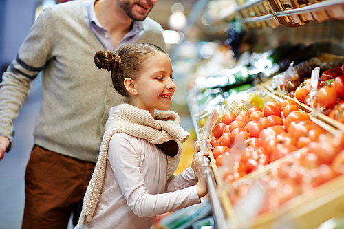 Smiling girl looking at red ripe tomatoes on supermarket shelf with her father near by