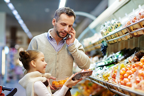 Portrait of man grocery shopping with daughter: speaking by phone calling wife to ask what vegetables to buy