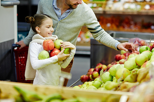 Girl holding heap of green and red apples while her father choosing them