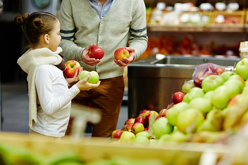 Portrait of cute little girl with dad choosing fresh ripe apples and other fruits in supermarket