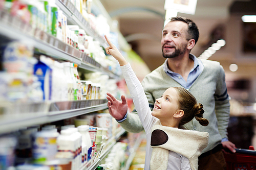 Happy girl pointing at pack of youghurt on upper shelf in supermarket