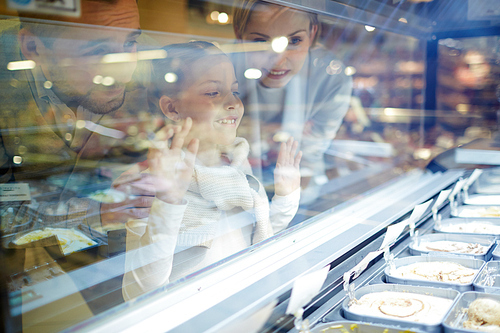 Portrait of happy family with cute little girl looking at ice cream through glass window in grocery store