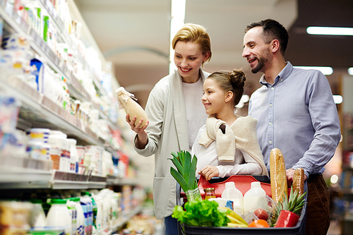 Portrait of happy family shopping in grocery store: choosing milk in dairy product department with cart full of food