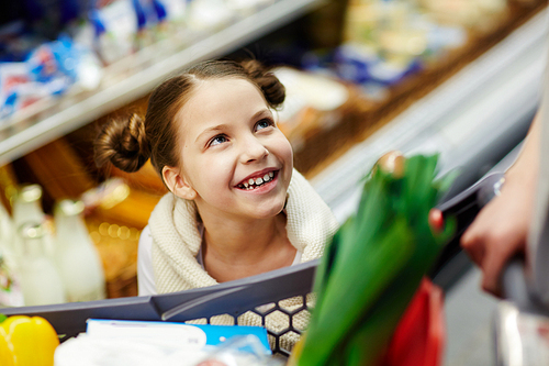 Portrait of cute little girl looking up at her parents from behind shopping cart while buying groceries in supermarket