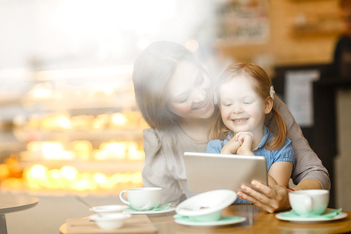 Woman showing her daughter interesting cartoon in touchpad while sitting in cafe