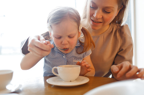 Adorable child sipping hot drink from spoon with her mother near by