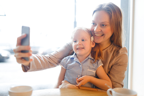 Happy modern female making selfie with her daughter in cafe