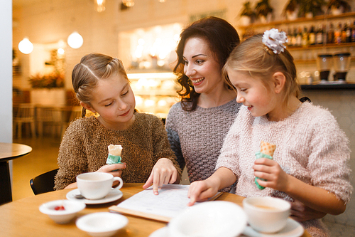 Twin girls with ice-cream pointing at document while talking to mother in cafe
