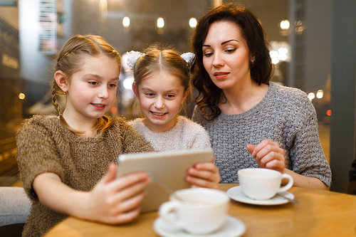 Twins and young woman looking at screen of touchpad by table in cafe