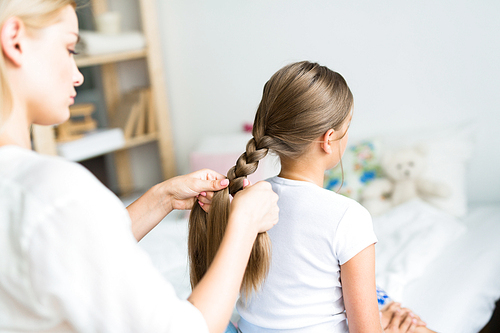 Young woman carefully braiding her daughters long beautiful hair sitting in sunlit childrens bedroom