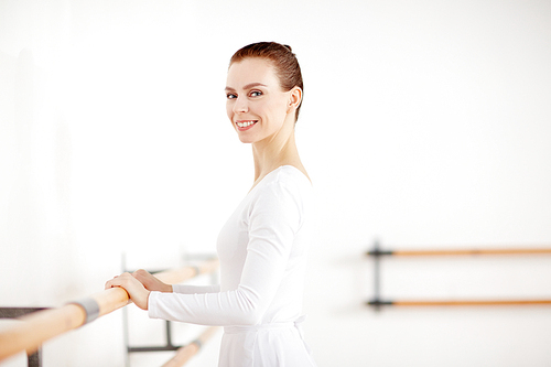 Happy young woman in ballet costume standing by wall of classroom and keeping her hands on wooden bar