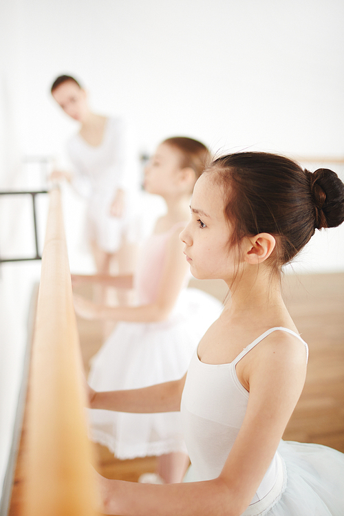 Little girls and her classmate in ballet outfit exercising by bar with teacher on background
