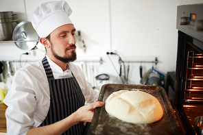 Young baker with tray putting raw loaf of bread into oven