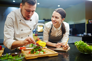 Young woman writing down recipe of vegetable salad while chief cooking it