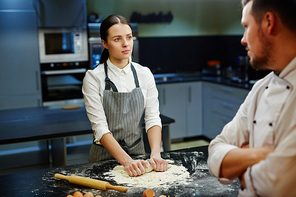 Serious young woman in apron kneading dough and looking at chef