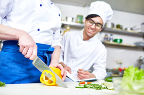 Trainee in uniform cutting pepper with chef near by