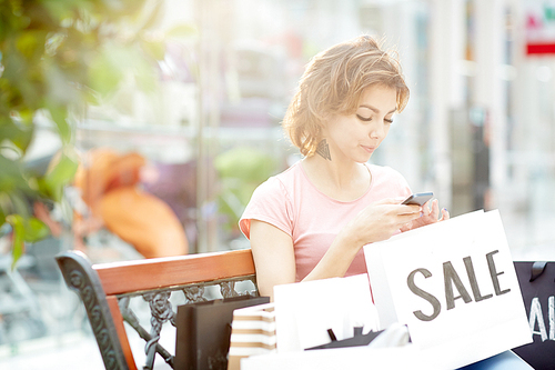 Young woman texting in smartphone while having rest after shopping