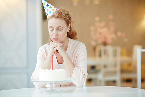 Sad woman in birthday cap sitting by table with dessert