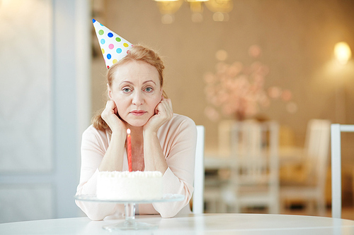 Portrait of sad  lonely mature woman sitting alone at birthday table with cake , wearing party hat and 